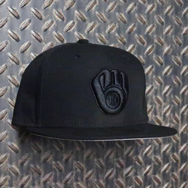 A black flat-brim baseball cap with a tonal black embroidered baseball glove logo on the front, displayed against a textured diamond plate metal background. New Era Milwaukee Brewers Ball & Glove 59FIFTY Fitted Hat Black 12344802