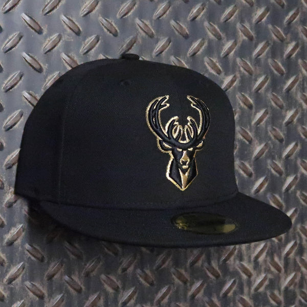 A black flat-brim baseball cap featuring an embroidered gold deer head logo on the front, displayed against a textured diamond plate metal background. New Era Milwaukee Bucks Black & Gold 59FIFTY Fitted Hat Black Gold 70686276