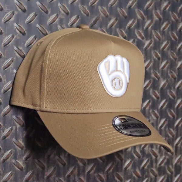 A tan baseball cap with a white embroidered Milwaukee Brewers glove logo on the front, resting on a textured diamond plate background. A black New Era 9FORTY sticker is visible on the brim. New Era Milwaukee Brewers Ball & Glove 9FORTY A-Frame Snapback Hat Khaki White 70776252