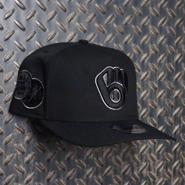 A black baseball cap with a black and white embroidered fist logo on the front and a circular patch on the side, displayed against a diamond plate metal background. New Era Milwaukee Brewers All-Star Game 1975 9FIFTY A-Frame Snapback Hat Black Silver 71083261