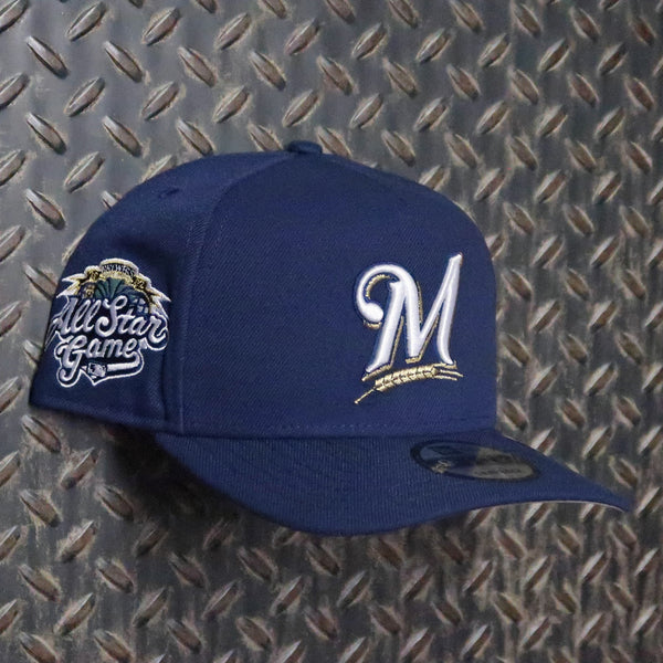 A navy blue Milwaukee Brewers baseball cap featuring a white embroidered 'M' logo on the front and an 'All Star Game' patch on the side, displayed against a textured diamond plate background. New Era Milwaukee Brewers All-Star Game 2002 9FIFTY A-Frame Snapback Hat Navy 71025030