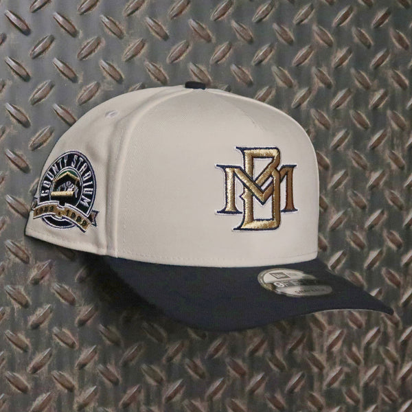 A baseball cap with an off-white crown, a dark navy blue visor, and embroidered details. The front features an embroidered 'MM' logo in gold and navy, and the side displays a circular 'Montana State Cavalry' patch. New Era Milwaukee Brewers County Stadium 9FIFTY A-Frame Snapback Hat 70872681