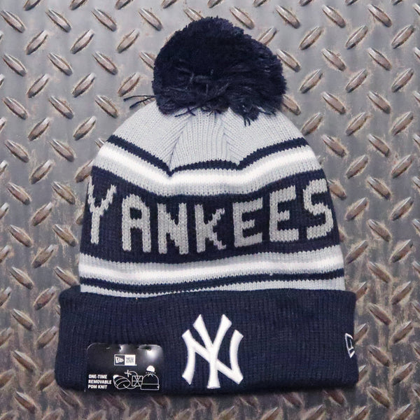 A navy blue and white knit beanie with a dark navy pom-pom, featuring the word 'YANKEES' across the front and a navy cuff with a white New York Yankees logo. The beanie is laid flat on a textured diamond plate background, with a hangtag still attached at the base. New Era New York Yankees Knit Pom Beanie Navy White 60399799