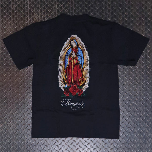 A black short-sleeve t-shirt laid flat on a textured silver diamond plate background, featuring a large graphic of Our Lady of Guadalupe on the back in red, blue, gold, and white, with the word 'Primitive' printed below in white script. Primitive Mission Heavyweight Knit T-Shirt Black PA425378