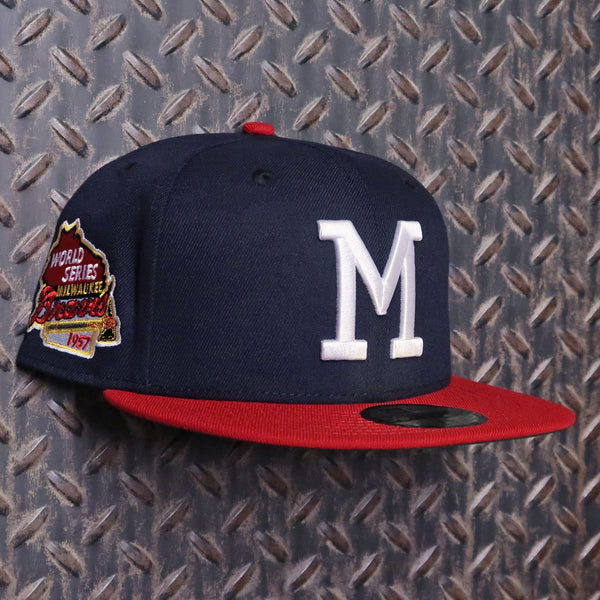 A navy blue baseball cap with a red flat brim, featuring a large white embroidered letter 'M' on the front and a World Series Champions 1977 patch on the side, set against a textured diamond plate background. New Era Milwaukee Braves World Series 2Tone 9FIFTY Snapback Hat Navy Red